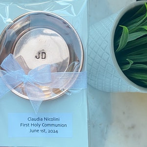 Puede incluir: Un plato redondo de color cobre con las iniciales "JD" estampadas en el centro, empaquetado con una cinta blanca. El plato est&aacute; empaquetado en pl&aacute;stico transparente. Una etiqueta dice "Claudia Nicolini Primera Comuni&oacute;n 1 de junio de 2024."