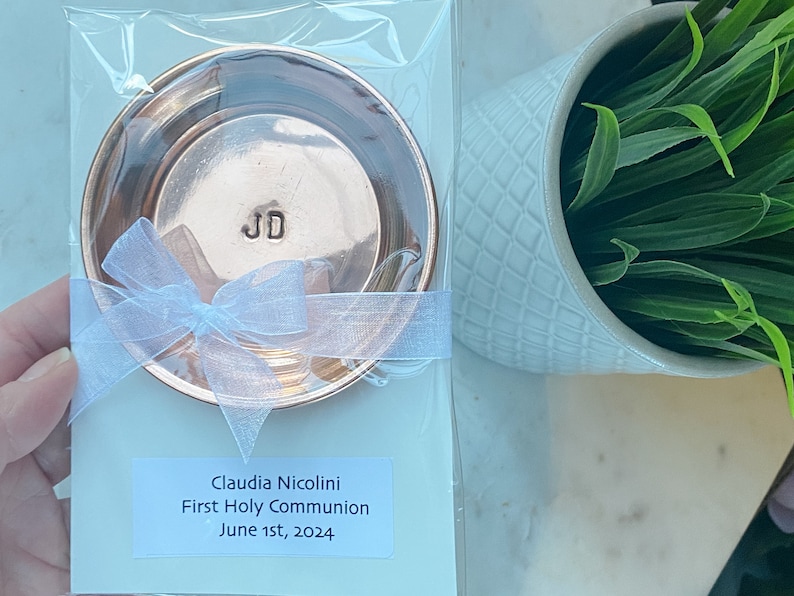 Puede incluir: Un plato redondo de color cobre con las iniciales "JD" estampadas en el centro, empaquetado con una cinta blanca. El plato est&aacute; empaquetado en pl&aacute;stico transparente. Una etiqueta dice "Claudia Nicolini Primera Comuni&oacute;n 1 de junio de 2024."