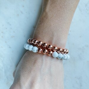 May include: Two bracelets stacked on a wrist. The top bracelet is made of copper-colored beads. The bottom bracelet is made of white and gray marble-like beads.