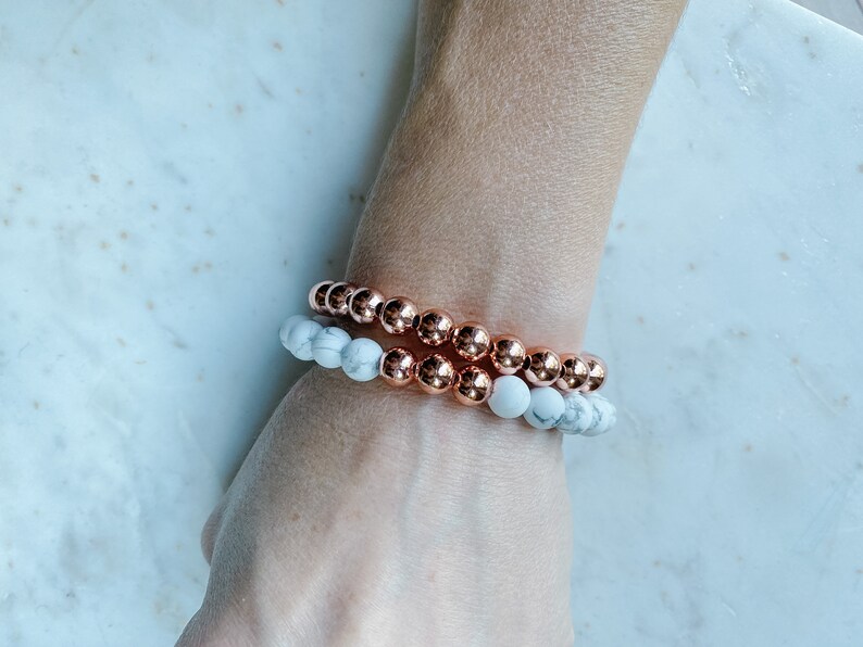 May include: Two bracelets stacked on a wrist. The top bracelet is made of copper-colored beads. The bottom bracelet is made of white and gray marble-like beads.