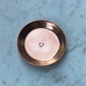 May include: A small, round copper dish with a heart stamped in the center. The dish has a slightly raised edge and a polished finish. The heart is a simple, minimalist design. The dish is likely used for holding small items like jewelry.