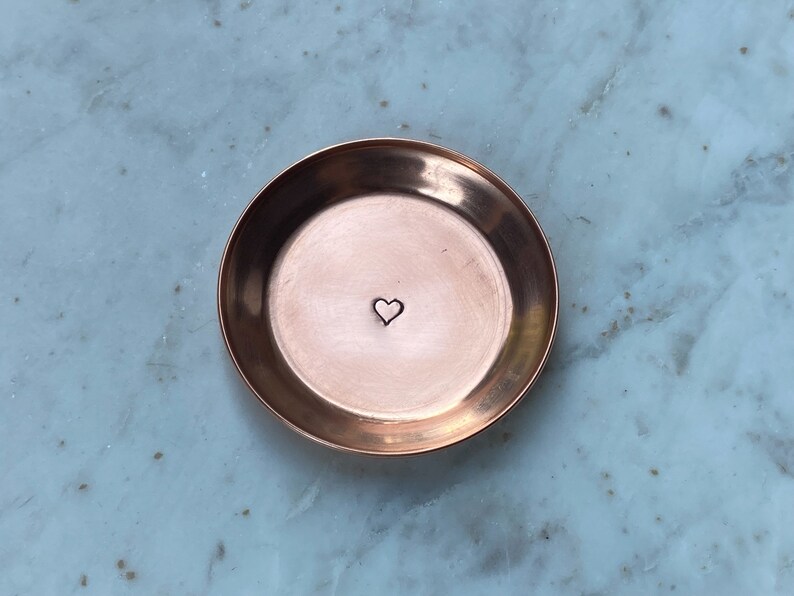 May include: A small, round copper dish with a heart stamped in the center. The dish has a slightly raised edge and a polished finish. The heart is a simple, minimalist design. The dish is likely used for holding small items like jewelry.