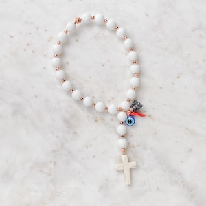 May include: White beaded bracelet with a red chilli pepper charm, a blue evil eye charm, and a white cross charm.