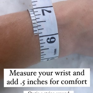 May include: A white measuring tape wrapped around a wrist to show how to measure for a bracelet. The text reads "Measure your wrist and add .5 inches for comfort. Or tie a string around your wrist and measure the string with a straight edge ruler."