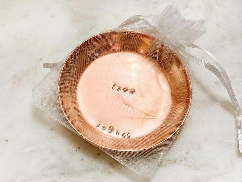 Puede incluir: Un peque&ntilde;o cuenco redondo de cobre con la palabra "love" y la fecha "9.9.06" grabadas. El cuenco est&aacute; dentro de una bolsa de organza blanca transparente. El cobre tiene un c&aacute;lido tono marr&oacute;n rojizo.