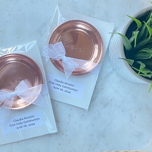 Puede incluir: Dos platos redondos de color cobre, cada uno empaquetado en pl&aacute;stico transparente con una cinta blanca. Cada plato tiene iniciales grabadas. El embalaje incluye una etiqueta que dice "Claudia Nicolini, Primera Comuni&oacute;n, 1 de junio de 2024."