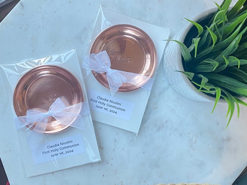 Puede incluir: Dos platos redondos de color cobre, cada uno empaquetado en pl&aacute;stico transparente con una cinta blanca. Cada plato tiene iniciales grabadas. El embalaje incluye una etiqueta que dice "Claudia Nicolini, Primera Comuni&oacute;n, 1 de junio de 2024."