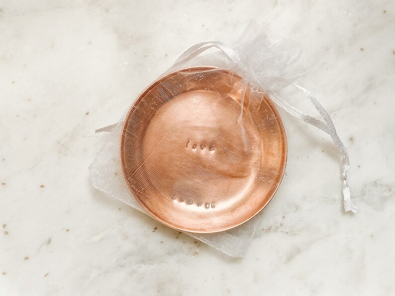 May include: A small, round copper dish with the word "love" stamped on it, presented in a sheer white gift bag. The dish has a textured surface and is likely used for holding small items. The word "love" is stamped in a simple, lowercase font.