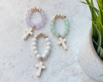 May include: Three beaded bracelets with cross pendants. One is lavender with "MILA", another is mint green with "LUCAS", and the third is white with "LOVED". Each has a white cross charm.