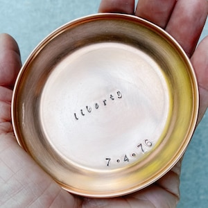 Puede incluir: Un peque&ntilde;o plato redondo de cobre con la palabra "liberty" y la fecha "7.4.76" estampadas en el metal. El plato tiene una superficie pulida y reflectante y se sostiene en una mano.