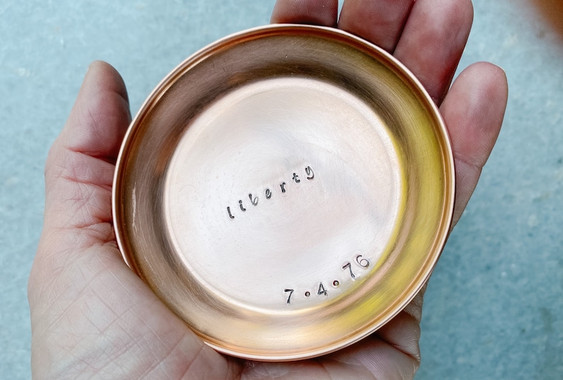 Puede incluir: Un peque&ntilde;o plato redondo de cobre con la palabra "liberty" y la fecha "7.4.76" estampadas en el metal. El plato tiene una superficie pulida y reflectante y se sostiene en una mano.