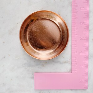 Puede incluir: Un peque&ntilde;o plato redondo de cobre, de aproximadamente 7.6 cm de di&aacute;metro. Una regla rosa se coloca junto al plato, mostrando medidas en pulgadas y cent&iacute;metros. El plato tiene una superficie reflectante y un borde elevado.