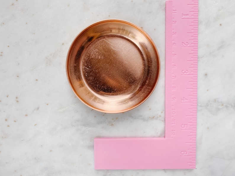 Puede incluir: Un peque&ntilde;o plato redondo de cobre, de aproximadamente 7.6 cm de di&aacute;metro. Una regla rosa se coloca junto al plato, mostrando medidas en pulgadas y cent&iacute;metros. El plato tiene una superficie reflectante y un borde elevado.