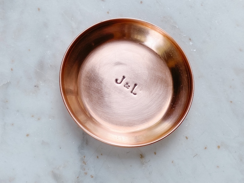 Puede incluir: Un peque&ntilde;o plato redondo de cobre con un borde ligeramente elevado. El interior del plato tiene las iniciales estampadas "J & L". El plato tiene una superficie pulida y reflectante y se encuentra sobre una superficie de m&aacute;rmol blanco y gris.