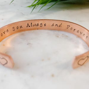 May include: A copper cuff bracelet with the inscription "Love you Always and Forever" engraved on the outside. The bracelet has two heart cutouts on the inside.
