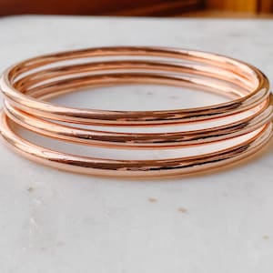 May include: Three rose gold bangle bracelets stacked on top of each other. The bracelets are smooth and shiny.