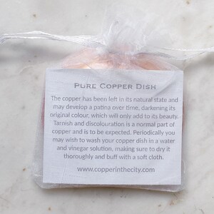 May include: A small, copper dish inside a sheer white drawstring bag. A white tag with black text reads "PURE COPPER DISH" and provides care instructions. The copper dish is in its natural state.