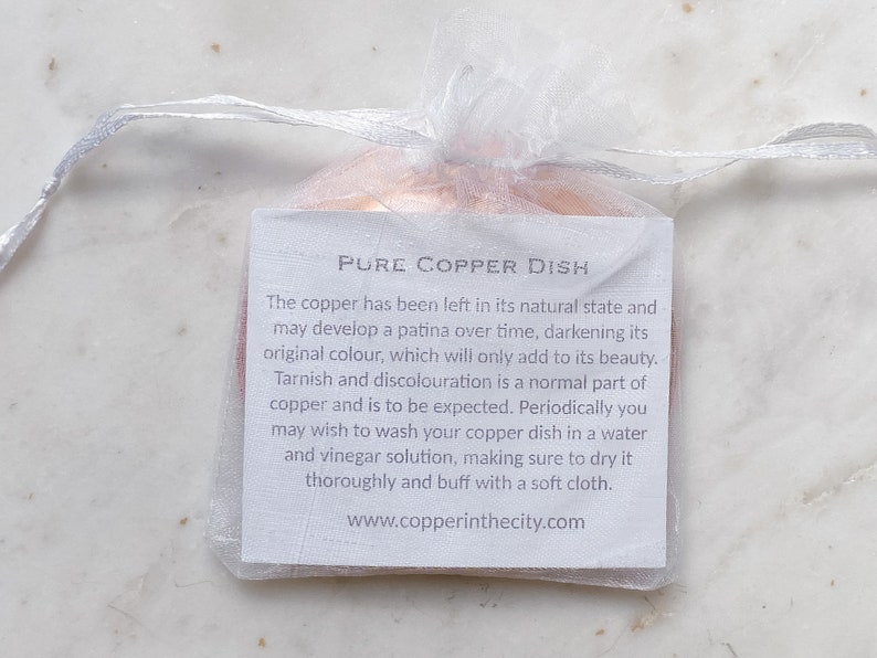 May include: A small, copper dish inside a sheer white drawstring bag. A white tag with black text reads "PURE COPPER DISH" and provides care instructions. The copper dish is in its natural state.