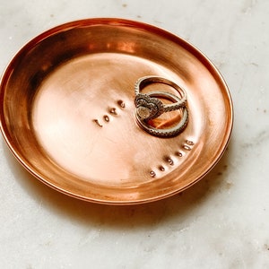 Puede incluir: Un peque&ntilde;o plato redondo de cobre con la palabra "love" estampada en la superficie. Dos anillos est&aacute;n colocados en el plato, y la fecha "9.9.06" est&aacute; estampada debajo. El plato tiene un dise&ntilde;o simple y elegante.