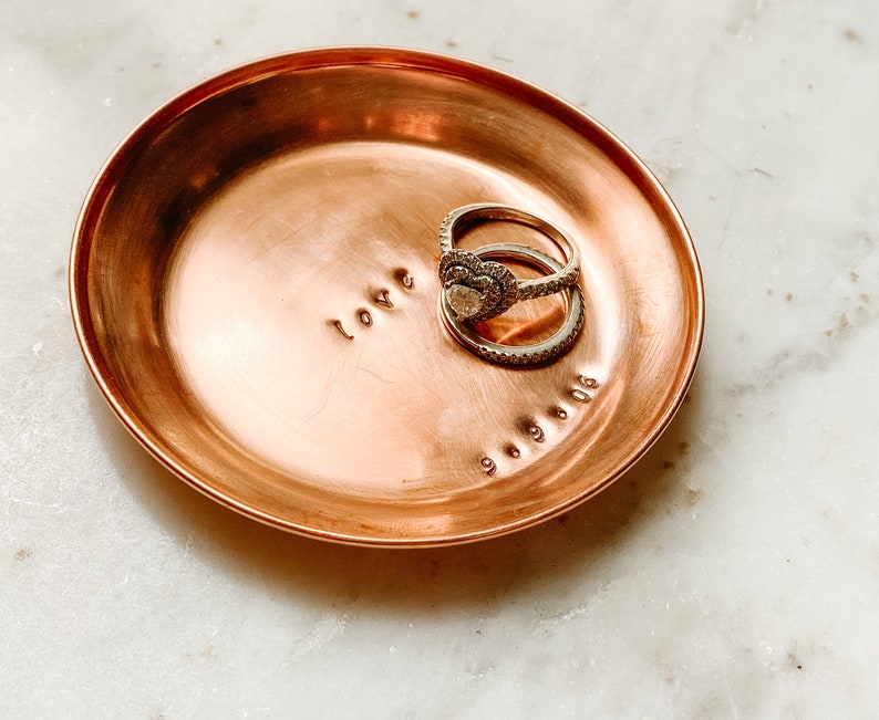 Puede incluir: Un peque&ntilde;o plato redondo de cobre con la palabra "love" estampada en la superficie. Dos anillos est&aacute;n colocados en el plato, y la fecha "9.9.06" est&aacute; estampada debajo. El plato tiene un dise&ntilde;o simple y elegante.