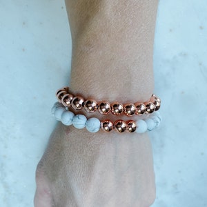 May include: Two bracelets on a wrist. The top bracelet is made of rose gold beads. The bottom bracelet is made of white beads.