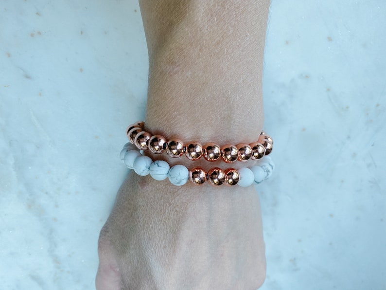 May include: Two bracelets on a wrist. The top bracelet is made of rose gold beads. The bottom bracelet is made of white beads.