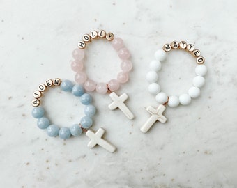 May include: Three beaded bracelets with gold letter beads spelling out the names Aiden, Holly, and Matteo. Each bracelet has a white cross charm.