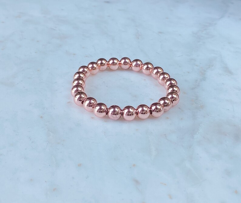May include: A rose gold beaded bracelet with a simple design. The bracelet is made of small, round beads that are strung together on a stretchy cord.