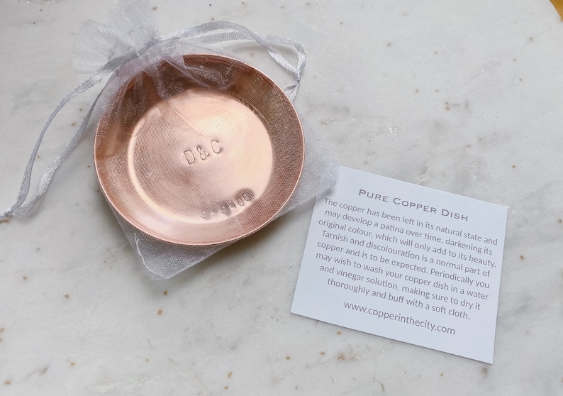 Puede incluir: Un peque&ntilde;o plato redondo de cobre con las iniciales "D&C" y la fecha "9-9-06" estampadas. El plato est&aacute; dentro de una bolsa transparente de color gris plateado. Una tarjeta blanca con texto est&aacute; al lado.