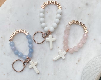May include: Three keychains with white cross charms and "blessed" spelled out in gold letters on the beads. The keychains are made with different coloured beads: white, light blue, and pink.