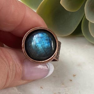 Peut inclure: Une bague de couleur cuivrée ornée d'une grosse pierre précieuse bleue irisée et ronde. La pierre précieuse présente un jeu de couleurs captivant, avec des éclats de bleu et de vert. La bande de la bague a un motif texturé.