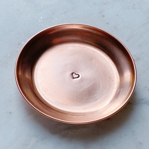 Puede incluir: Un peque&ntilde;o plato redondo de cobre con una hendidura en forma de coraz&oacute;n en el centro. El plato tiene una superficie lisa y pulida y un borde ligeramente elevado. El cobre tiene un tono marr&oacute;n rojizo c&aacute;lido. Este plato decorativo es ideal para objetos peque&ntilde;os.