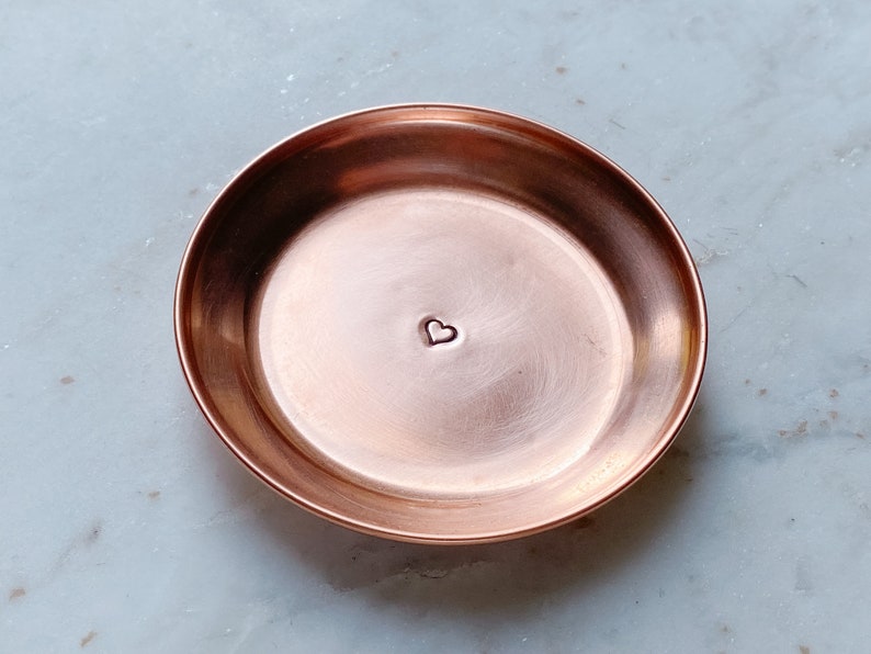 Puede incluir: Un peque&ntilde;o plato redondo de cobre con una hendidura en forma de coraz&oacute;n en el centro. El plato tiene una superficie lisa y pulida y un borde ligeramente elevado. El cobre tiene un tono marr&oacute;n rojizo c&aacute;lido. Este plato decorativo es ideal para objetos peque&ntilde;os.