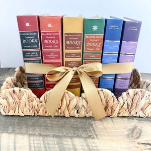 May include: A collection of vintage Reader's Digest Condensed Books in a woven basket, tied with a gold ribbon. The books have various colored spines, including red, orange, yellow, green, blue, and purple. The books are arranged in a decorative display.