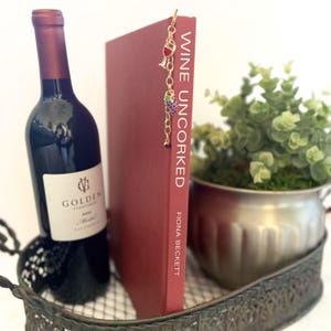 May include: A bottle of red wine next to a burgundy book with the title "Wine Uncorked" and a gold bookmark with wine glass and grape charms. The book and wine are in a decorative metal basket.