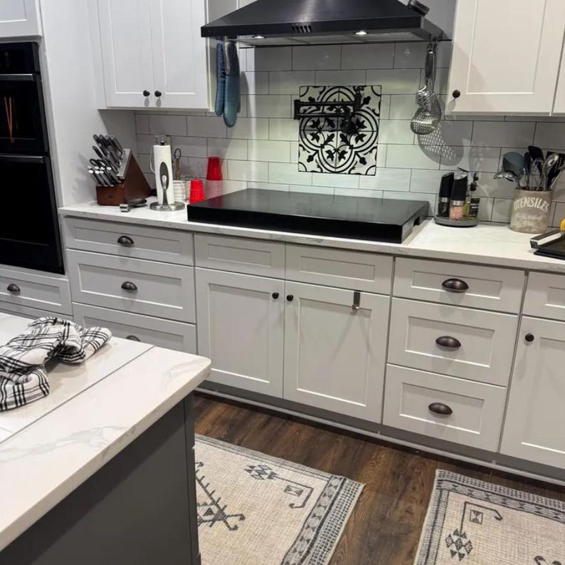 May include: White kitchen cabinets with black hardware and a black electric stovetop. The backsplash is white subway tile with a black and white patterned tile accent. The countertop is white marble.