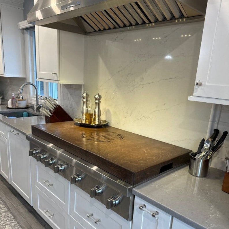 May include: A stainless steel stovetop with a wooden cutting board on top. The cutting board has a decorative design and the words "Gather Here" engraved on it. The stovetop is in a modern kitchen with white cabinets and a gray countertop.