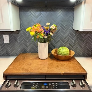 May include: A wooden stovetop cover with a light brown wood grain finish. The cover is placed over a black stovetop with a digital display and knobs. A vase with a white textured finish holds a bouquet of colorful flowers. A wooden bowl with a watermelon inside sits on the cover.