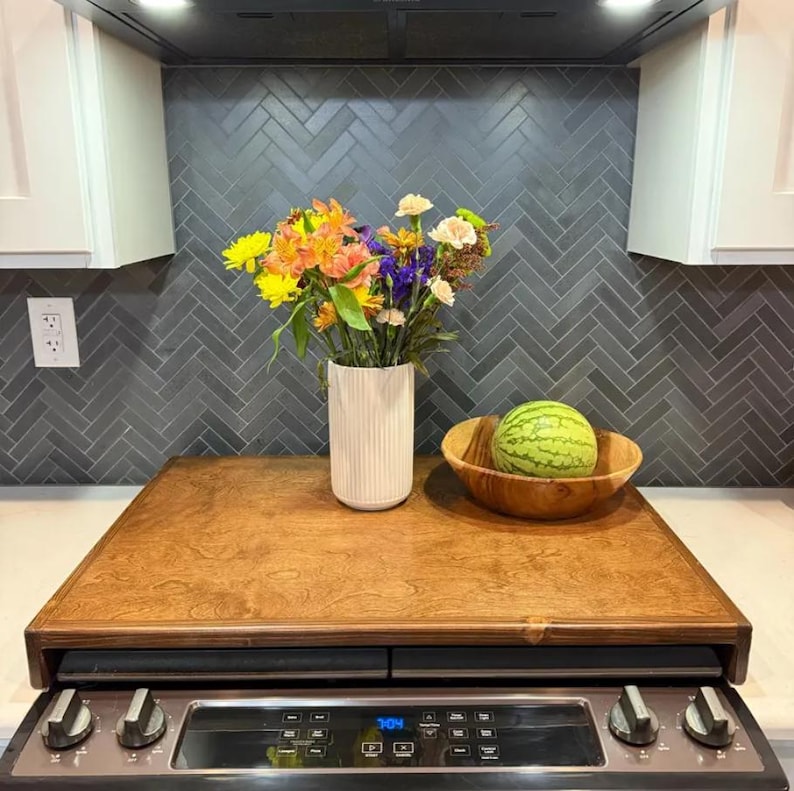 May include: A wooden stovetop cover with a light brown wood grain finish. The cover is placed over a black stovetop with a digital display and knobs. A vase with a white textured finish holds a bouquet of colorful flowers. A wooden bowl with a watermelon inside sits on the cover.