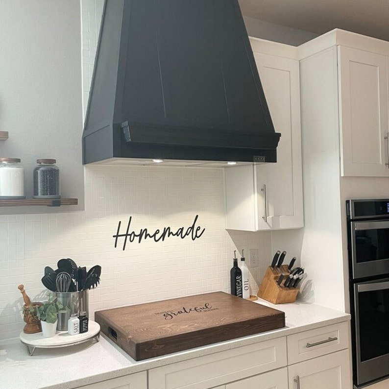May include: A dark wood cutting board with the words "Homemade with love" and "Grateful" engraved on it. The board is sitting on a white countertop in a kitchen with white cabinets and a black range hood.