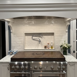 May include: A stainless steel stove with a wooden cutting board on top. The stove has six burners and a double oven. The cutting board has a decorative design in the center.