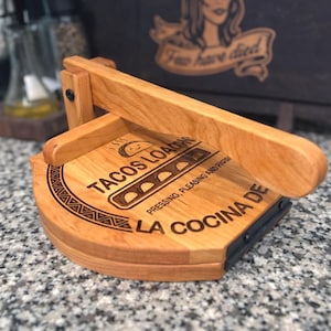 May include: Wooden tortilla press with a hinged design. The press is light brown with engraved text that reads "TACOS LOADING" and "LA COCINA DE". The press is on a speckled gray surface.