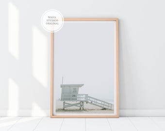 May include: A framed print featuring a lifeguard station on a sandy beach. The lifeguard station is painted a light blue and has the number 26 on it. The print is in a light wood frame and has the text "MAHA STUDIOS ORIGINAL" in the upper left corner.