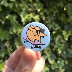 May include: A blue and white button featuring a cartoon illustration of a dog wearing sunglasses and skateboarding. The dog is brown and has a pink skateboard.
