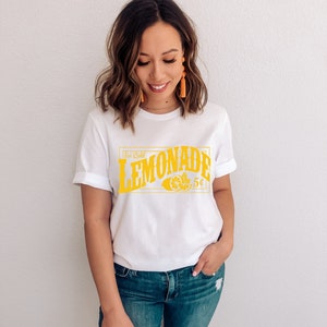 May include: A white t-shirt with a yellow graphic that says "Ice Cold Lemonade 5&cent;". The graphic features a lemon and a leaf.