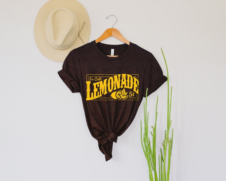 May include: A brown t-shirt with a yellow graphic that says "Ice Cold Lemonade 5&cent;" with a lemon illustration.