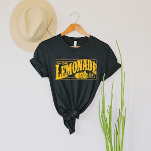 May include: A dark grey t-shirt with a yellow graphic that says "Ice Cold Lemonade 5&cent;" with a lemon slice illustration.