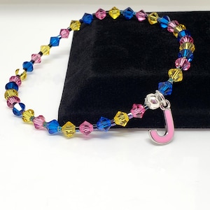 May include: A beaded bracelet featuring faceted bicone crystals in alternating shades of blue, yellow, and pink. The bracelet includes a silver-toned metal charm in the shape of the capital letter J, which has a smooth, light pink enamel finish.