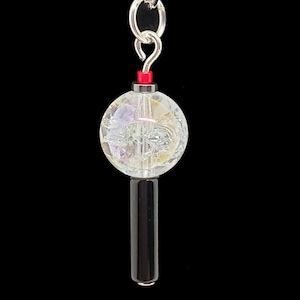 May include: A keychain charm featuring a clear, crackled glass sphere with internal purple and yellow inclusions, topped with a small red bead and a silver-toned metal jump ring, attached to a smooth, cylindrical black bead at the base.
