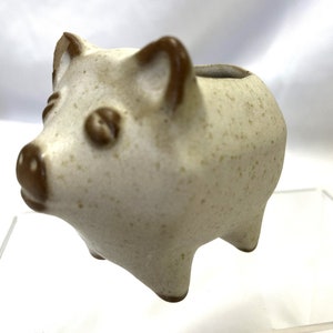 May include: A small white ceramic piggy bank with brown ears and snout. The piggy bank has a speckled glaze and is standing on a clear surface.